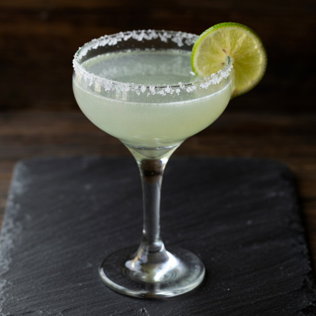 Silver edible glitter margarita in a coupe glass with salted rim and lime wheel, silver luster dust particles catching moody low light